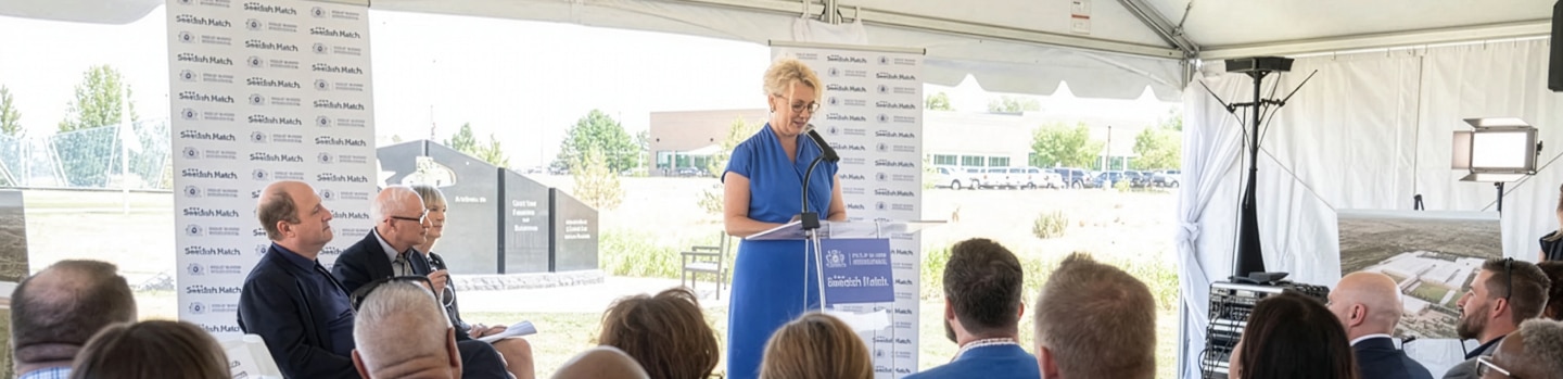 A speaker in a blue dress addresses an audience under a tent, with several attendees seated and listening attentively.